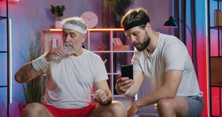Likable smiling senior sporty man sitting on fitball and drinking water simultaneously watching funny video on phone in his grandson's hands while they relaxing between sports training at home