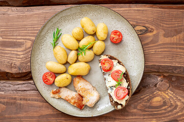 Boiled young potatoes, chunks of grilled turkey meat and cereal bread with blue cheese and tomatoes on a plate on a wooden background.