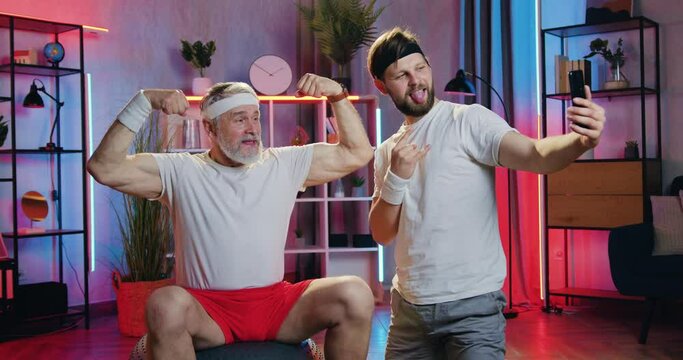 Attractive smiling bearded old man showing his muscles and likable funny young guy making joint selfie with grimaces during evening home workout