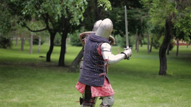 Battle of two medieval warriors in full sets of armor with swords and shields. Duel of knights in the park.