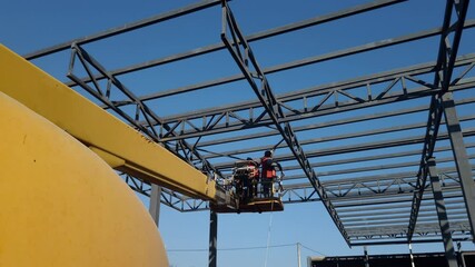 Worker on a cherry picker, together with telescopic crane, installs a concrete block. It is part of an industrial building metal frame under construction