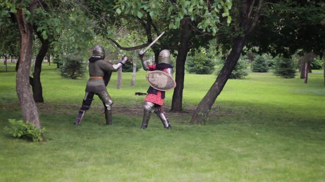 Battle of two medieval warriors in full sets of armor with swords and shields. Duel of knights in the park.