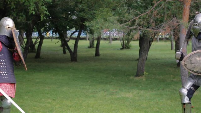 Battle of two medieval warriors in full sets of armor with swords and shields. Duel of knights in the park.