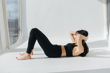 young armenian woman in black sportswear practicing yoga on white floor near window