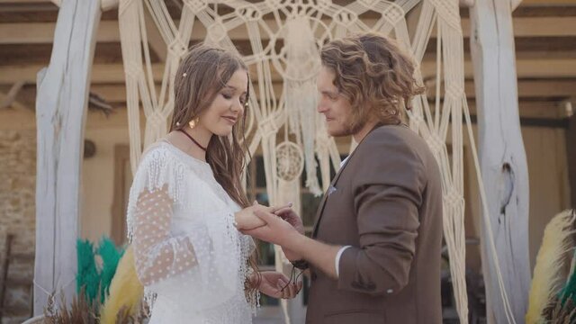 Wedding in hippie style. Action. Beautiful couple of newlyweds will exchange rings under crown. Hippie newlyweds get engaged in beautiful outfits on background of braided crown