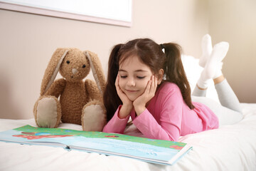 Little girl with toy bunny reading book on bed at home
