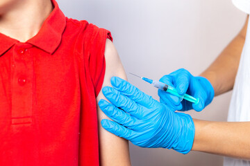 A nurse in a white cap, disposable mask and blue gloves injects the vaccine through a syringe into the shoulder of a brave boy in a red T-shirt.
