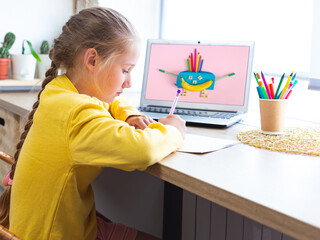 Caucasian schoolgirl does homework, draws in a notebook. Cozy workplace by the window. Multicolored school supplies, office supplies and a banana in the form of a smiling character on a laptop screen.