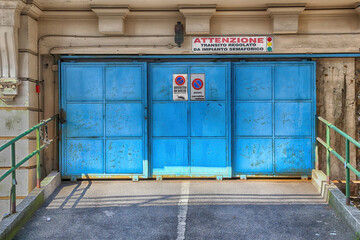 Old blue closed door of a Garage in Italy. Warning sign indicating transit regulated by a traffic light system and no parking sign attached at the door.