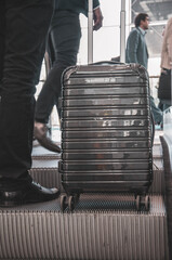 Stylish grey suitcase with wheels on escalator moving stairs near businessman leg in expensive suit and shoes. Business trip concept. Closeup reflective surface of modern hand luggage in airport