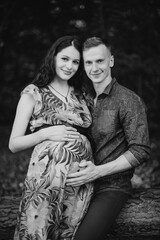 Black and white photo. Close up portrait of happy pregnant couple, standing in beautiful summer forest, touching belly and smiling at camera