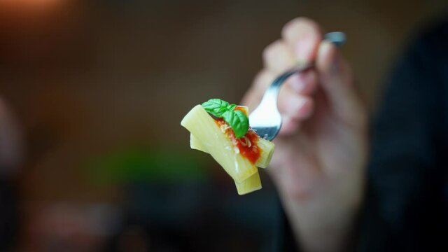 Man holds a fork with two delicious Italian penne pasta macaroni with tomato sauce and basil and brings it closer to the camera.