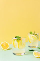 Two glasses of ice lemon water with mint leaves on a yellow and green background. Fresh summer drink with soda.