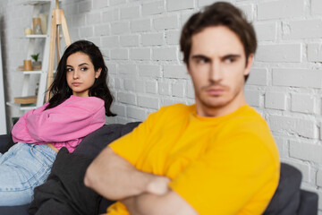 young woman sitting on couch near blurred disappointed man with crossed arms at home