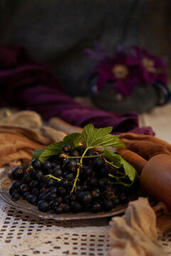 Black currant berries  with leaves in metal plate on white  embroidered tablecloth  with potato masher and violate flowers, selective focus, fresh juice making concept, dark background, vertical