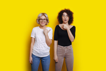 Obraz premium Adorable women with curly hair are smiling at camera and pointing at each other on a yellow studio wall
