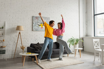 smiling young couple in casual clothes dancing in living room
