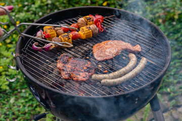 Smoking charcoal grill with barbecue tongs. Grill grate with steaks, sausages and vegetable skewers.