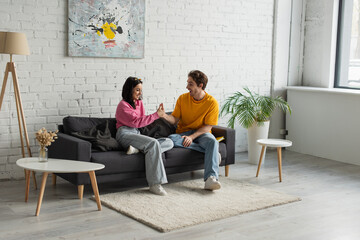 Fototapeta premium playful young couple sitting on couch and touching with hands in living room