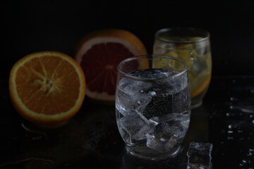 Fototapeta na wymiar splashes of water over a glass of water and ice. Nearby are fruits orange and grapefruit, ice. On a black background. Refreshing cold ice drink