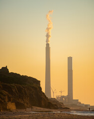 A power plant with chimneys emitting smoke by the sea and silhouettes of people nearby on the beach and on a cliff.
