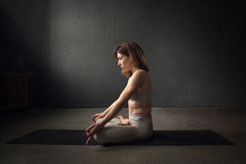 Fototapeta na wymiar A slim beautifully lighted young woman is doing sukhasana yoga pose in a dark room. Image with selective focus and toning
