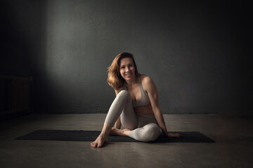 Fototapeta na wymiar A slim beautifully lighted young woman is doing yoga in a dark room. Image with selective focus and toning