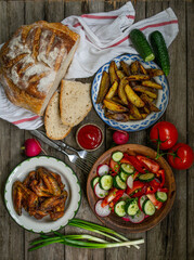 French fries, wings, salad, vegetables, bread on an old wooden background. Rural dinner, summer picnic. Top view. Flat lay.