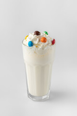 glass with white milkshake and cream decorated with colored candies on a gray background with...