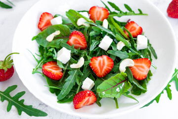 Tasty salad of strawberry with arugula, young beet tops, feta cheese and cashew nuts in white plate on marble table with salad leaves and fresh strawberry fruits around. Fruit detox and antioxidants.