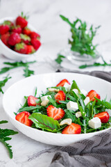 Summer salad of strawberry with arugula, spinach leaves, feta cheese and cashew nuts in white plate with gray towel, salad leaves and fresh strawberry fruits around. Easy recipe for everyday.