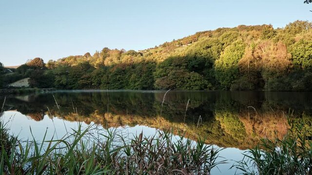 A beautifully calm sunrise time lapse over Sparth Reservoir, West Yorkshire, UK. The glassy reservoir shows a beautiful colourful reflection of the sun shining on the trees on the opposite side as the