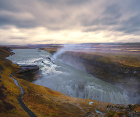 The Gullfoss waterfall in Iceland shimmers in the evening sunset under a cloudy sky.