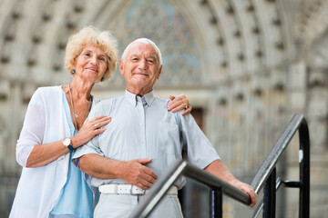 Romantic glad cheerful positive senior couple standing near iron railings in front of cathedral