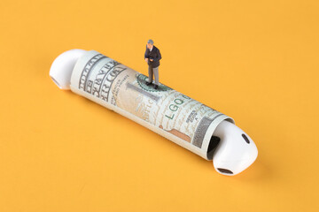 Miniature figures stand on a roll of dollar bills with headphones inserted at both ends of the roll