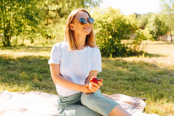 Portrait of a happy young woman with glasses enjoying summer time, outdoors. Beautiful blonde girl holding a fruit and having a rest in the nature outside. Summer lifestyle