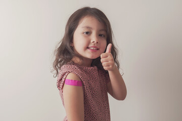 Young mixed Asian girl giving thumb up and showing her arm with pink bandage after got vaccinated...