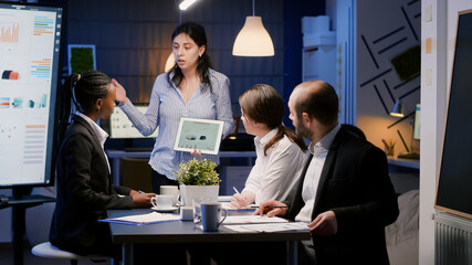 Fototapeta premium Business entrepreneur woman presenting management graphs using tablet working at company solution late at night in office meeting room. Diverse multi-ethnic teamwork brainstorming financial strategy