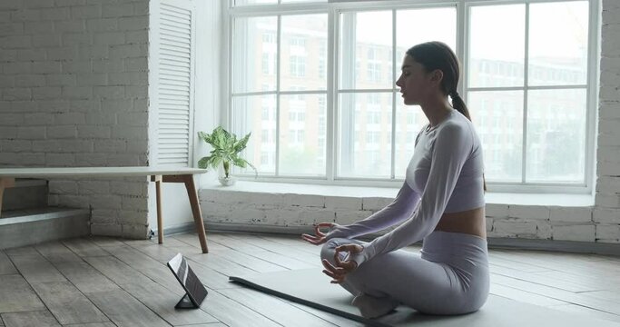 Young happy woman practicing yoga and doing breathing exercises watching live video learning classes on tablet. Female brunette enjoying calm relax harmony Zen mindfulness meditation at home