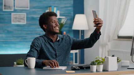 Authentic happy african american man smiling and taking a selfie in the living room to share it on social media. Mobile photo, online internet web leisure activity for black guy