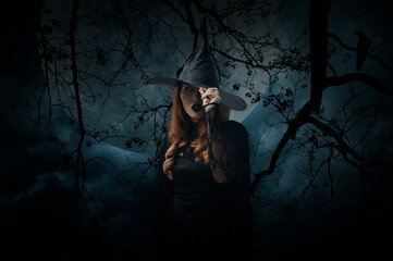 Scary halloween witch standing over dead tree, crow, birds, full moon and spooky cloudy sky,...