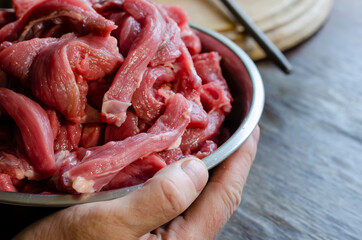 A man's hands hold a full metal bowl of sliced beef.