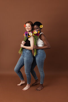 Two women with different skin color posing against a brown background. Cheerful females with flowers standing together looking at camera.