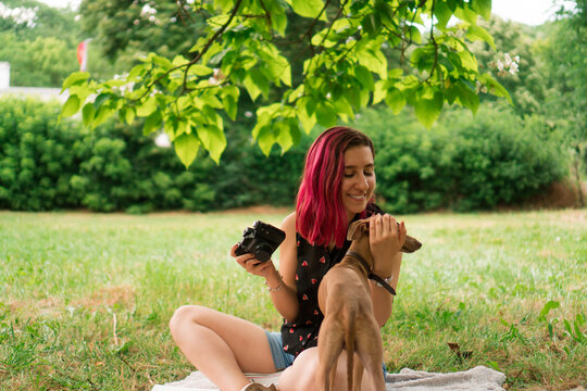 A young woman and her dog are sitting on the blanket in the park. She is taking photos with her vintage camera.