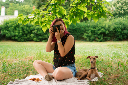 A young woman and her dog are sitting on the blanket in the park. She is taking photos with her vintage camera.