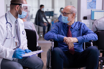 Paralized old man in wheelchair with protection mask listening doctor sitting in waiting area of...