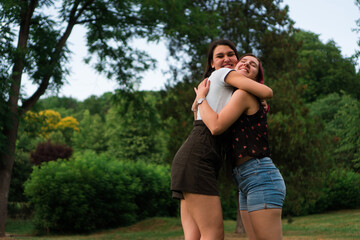 Two young women are hugging in the park, surrounded by beautiful greenery. They are smiling from ear to ear.