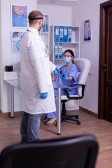 Specialist doctor wearing protection visor taking radiography from medical assistant to examinate next patient. Nurse with face mask against covid-19 sitting on chair working at pc in waiting area