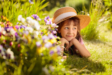 Happy little girl in a straw hat dreams while lying on the grass in the garden.