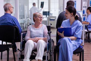 Nurse filing documents while talking with disabled senior woman sitting in wheelchair in hospital waiting area. Elderly paralyzed patient explaning symptoms, assitant checking registration form